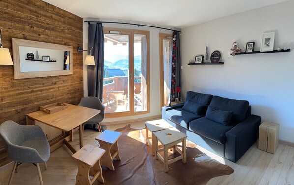 A cozy living room with a wooden wall, a dark blue sofa, a wooden dining table with chairs, and a small wooden stool. - Studio Charmant Front de Neige (Huez)