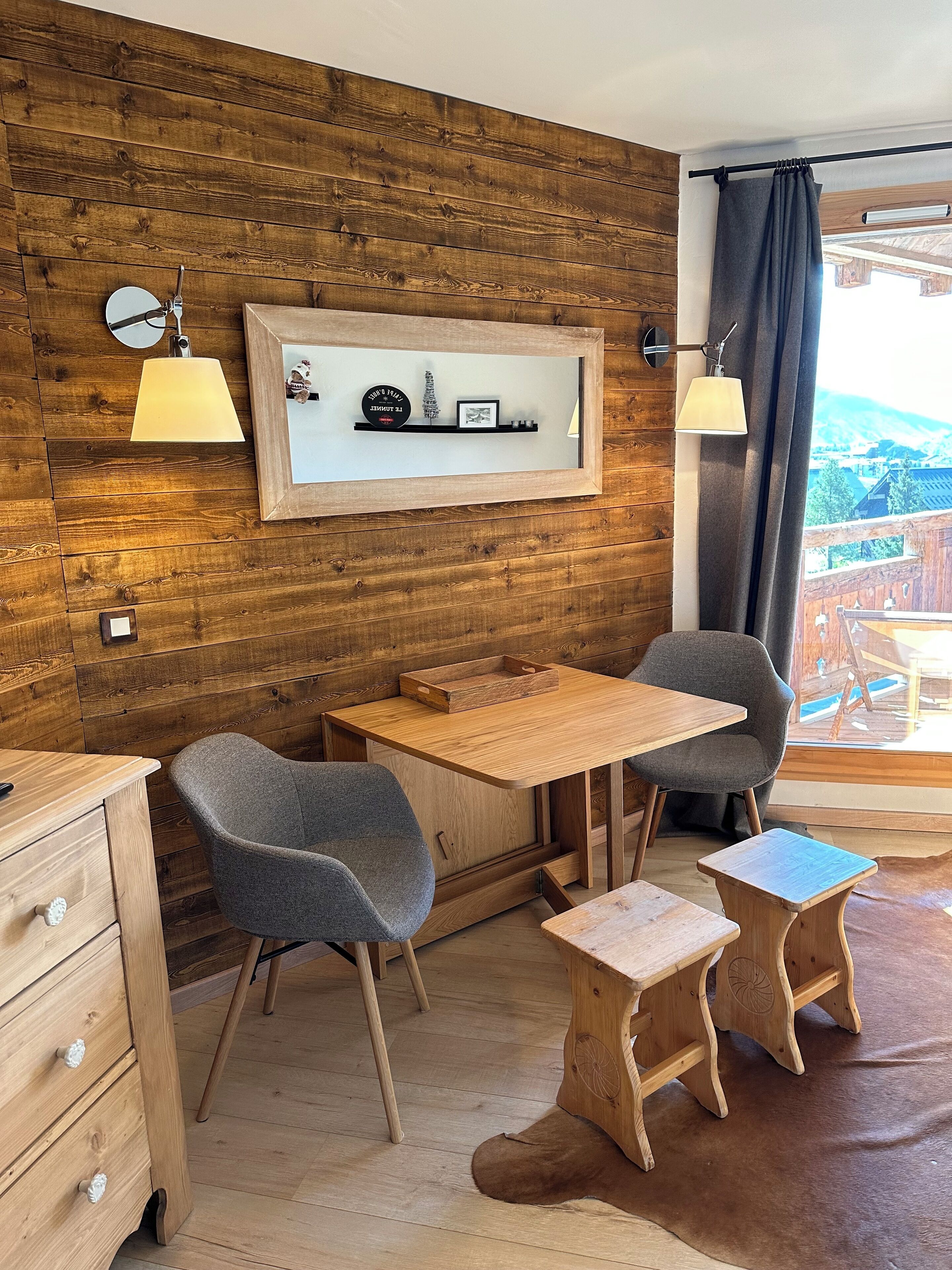 A cozy room with wooden walls, a wooden table, two chairs, a small cabinet, and a shelf with decorative items.