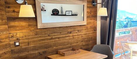 A cozy room with wooden walls, a wooden table, two chairs, a small cabinet, and a shelf with decorative items.