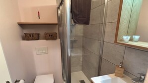 Moderni kylpyhuone, jossa on lasiseinällinen suihku, valkoinen wc-istuin ja peilillä varustettu pesuallas.