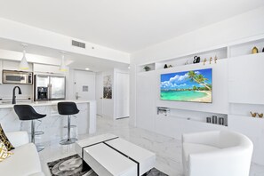 Et moderne køkken med hvide skabe, en mikroovn og et køleskab. Der er to sorte barstole og et hvidt spisebord. Et stort TV er monteret på væggen over en hvid sofa.