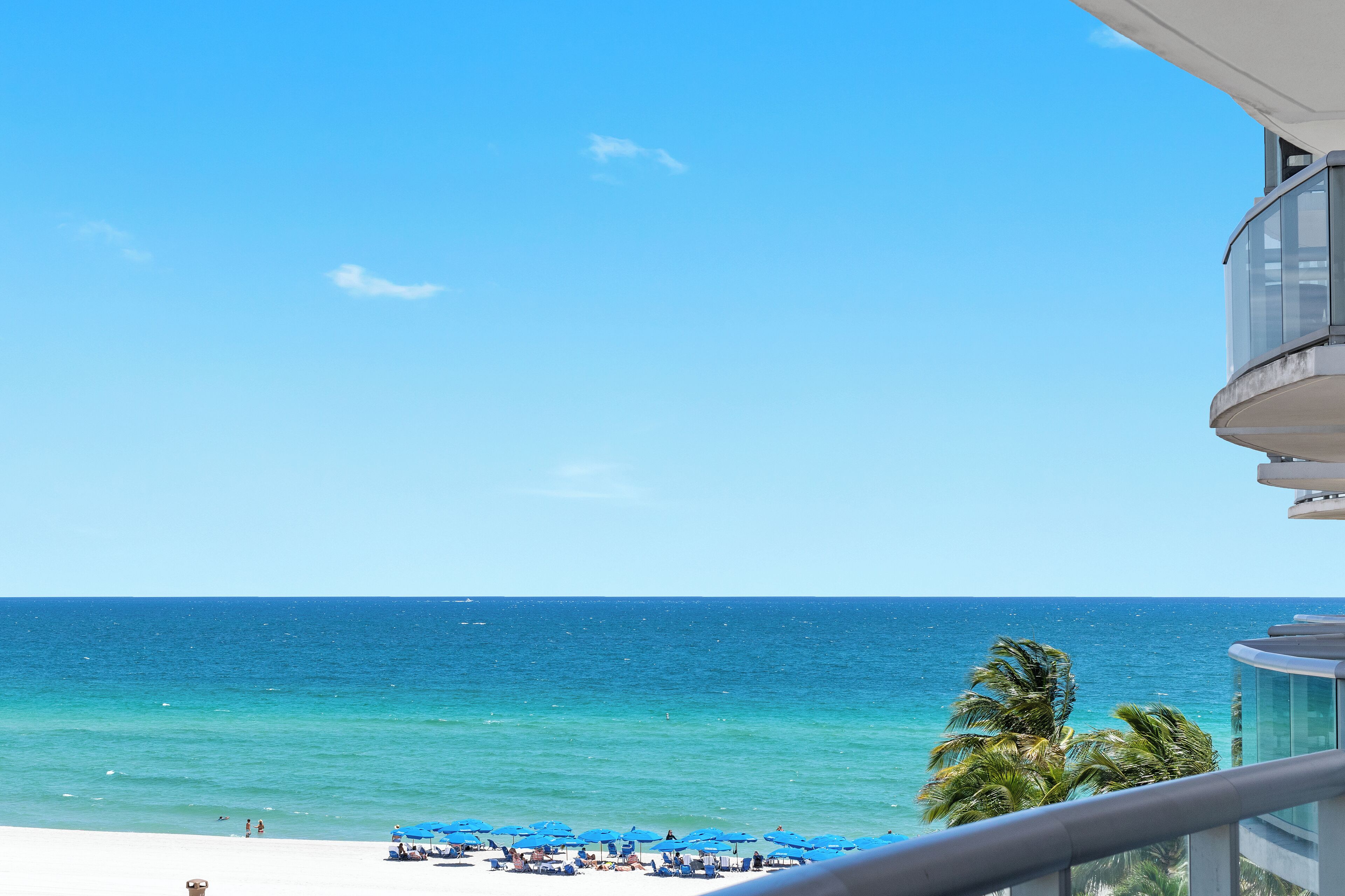 A beach view from a balcony with clear skies, ocean, and palm trees.