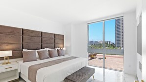 Un dormitorio moderno con una cama grande, un banco, una mesita de noche y vistas a una obra en construcción a través de un ventanal.