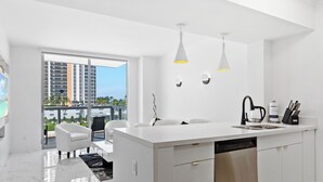 Una cocina moderna con una isla grande, armarios blancos y lavavajillas. Hay un comedor con una silla blanca y vistas a la ciudad a través de ventanas amplias.