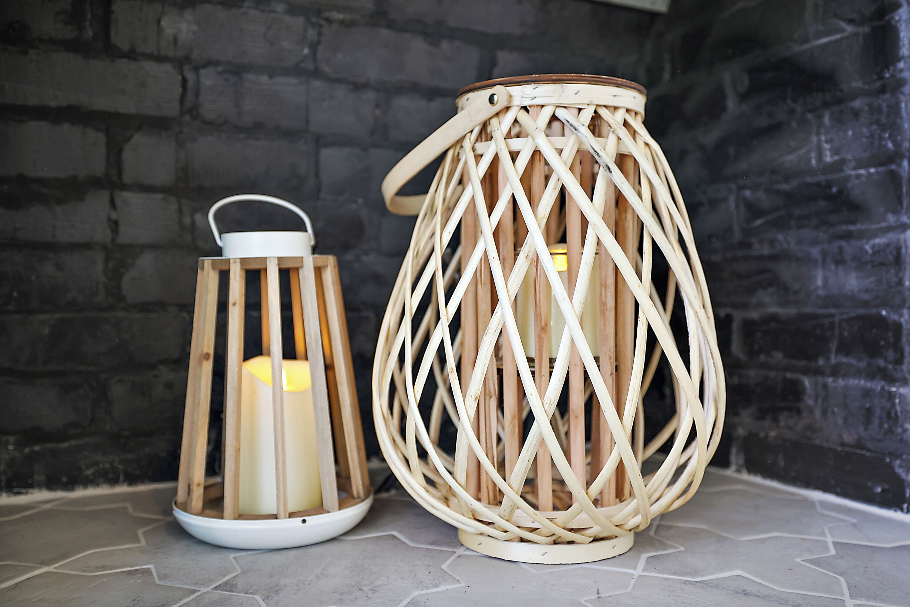 Deux lanternes en osier, avec des bougies à l’intérieur, sont posées sur un sol carrelé, adossées à un mur de briques.