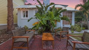 A patio with wicker furniture and a wooden table, surrounded by plants and a house with a white exterior.