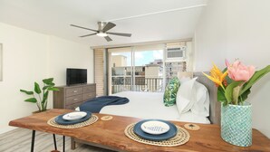 Ein modernes Schlafzimmer mit Bett, einem Holztisch mit Tellern, einem Fernseher, einem Deckenventilator, einem Balkon mit Blick und einer Vase mit Blumen.