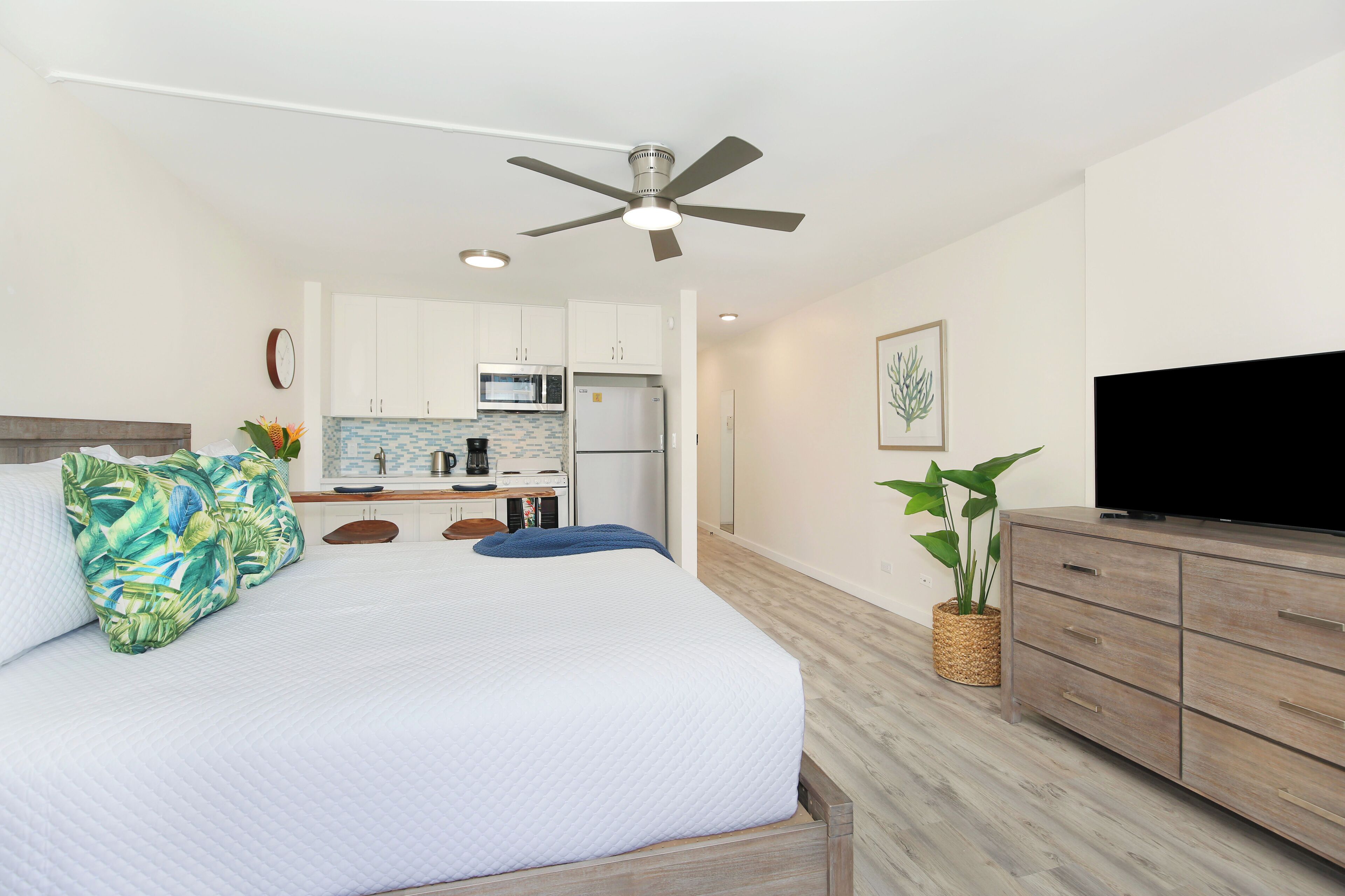 Ein Schlafzimmer mit Bett, Fernseher, Deckenventilator, Küchenzeile und einer Zimmerpflanze.