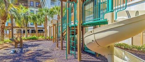 A playground with slides and climbing structures, surrounded by palm trees and a building with balconies.