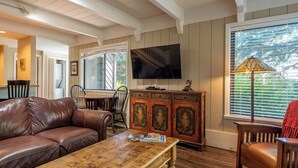 Una sala de estar con un sofá de cuero marrón, una mesa de centro de madera, un televisor montado en la pared, un mueble de madera y una ventana con persianas.