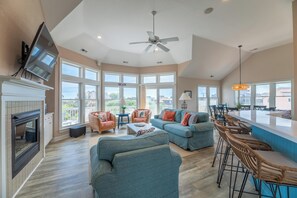 A spacious living area with a fireplace, comfortable seating, a large window with a view, and a ceiling fan. - Make Way For Ducklings - Wild Duck Dunes, Ocean Views, 160 Yds to Beach, Saltwater Pool, Flex Stays (Duck)