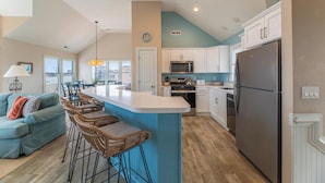 A modern kitchen with a central island, white cabinets, stainless steel appliances, and a dining area with a blue sofa and wicker bar stools. - Make Way For Ducklings - Wild Duck Dunes, Ocean Views, 160 Yds to Beach, Saltwater Pool, Flex Stays (Duck)