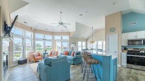 A spacious living area with a large window, a ceiling fan, and a kitchen with modern appliances. - Make Way For Ducklings - Wild Duck Dunes, Ocean Views, 160 Yds to Beach, Saltwater Pool, Flex Stays (Duck)