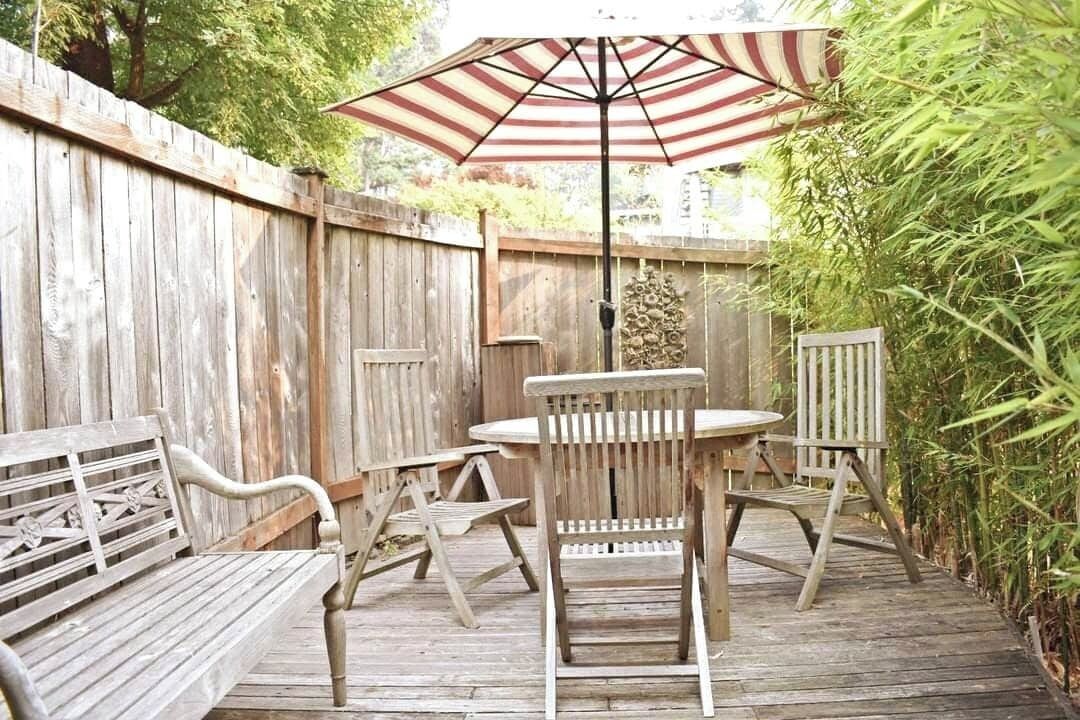 En udendørs patio med træterrasse, et rundt bord, to stole og en stribet parasol.