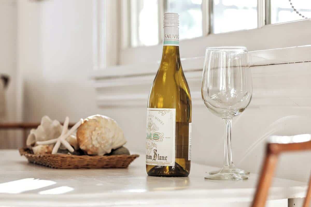 A bottle of white wine and an empty wine glass on a table with a basket of bread and cheese.