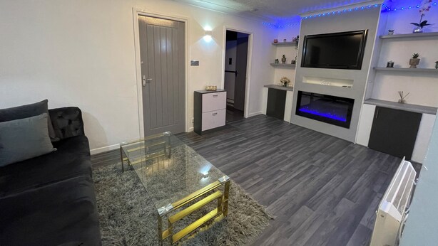 A modern living room with a glass coffee table, dark sofa, and a flat-screen TV mounted on the wall. - Newly Refurbished Modern 2 Bedroom Flat (Trimley Saint Mary)