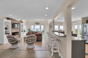 A spacious living area with a stone fireplace, white wicker chairs, a sofa with colorful cushions, and a view of the ocean through large windows. - Topsail's Treasure #148 - Oceanfront Home w/ Community Pool & Sound Access! (North Topsail Beach)