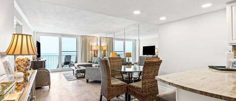 Une chambre d’hôtel moderne dotée d’un coin repas, d’un salon avec un canapé et des fauteuils, d’un coin cuisine et de grandes fenêtres offrant une vue panoramique.