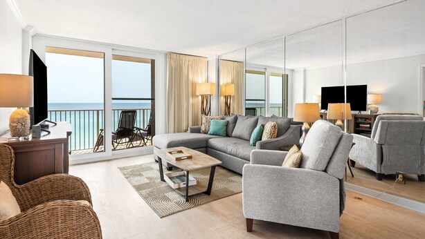 Een moderne woonkamer met een grote bank, fauteuils, een salontafel en een televisie. Er is een balkon met uitzicht op de oceaan.