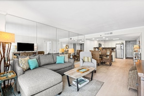 A modern living room with a sectional sofa, a coffee table, and a dining area with wicker furniture. - Gulf Front with Beach Setup, Pool, Private Beach + Free Attraction Tickets! (Santa Rosa Beach)