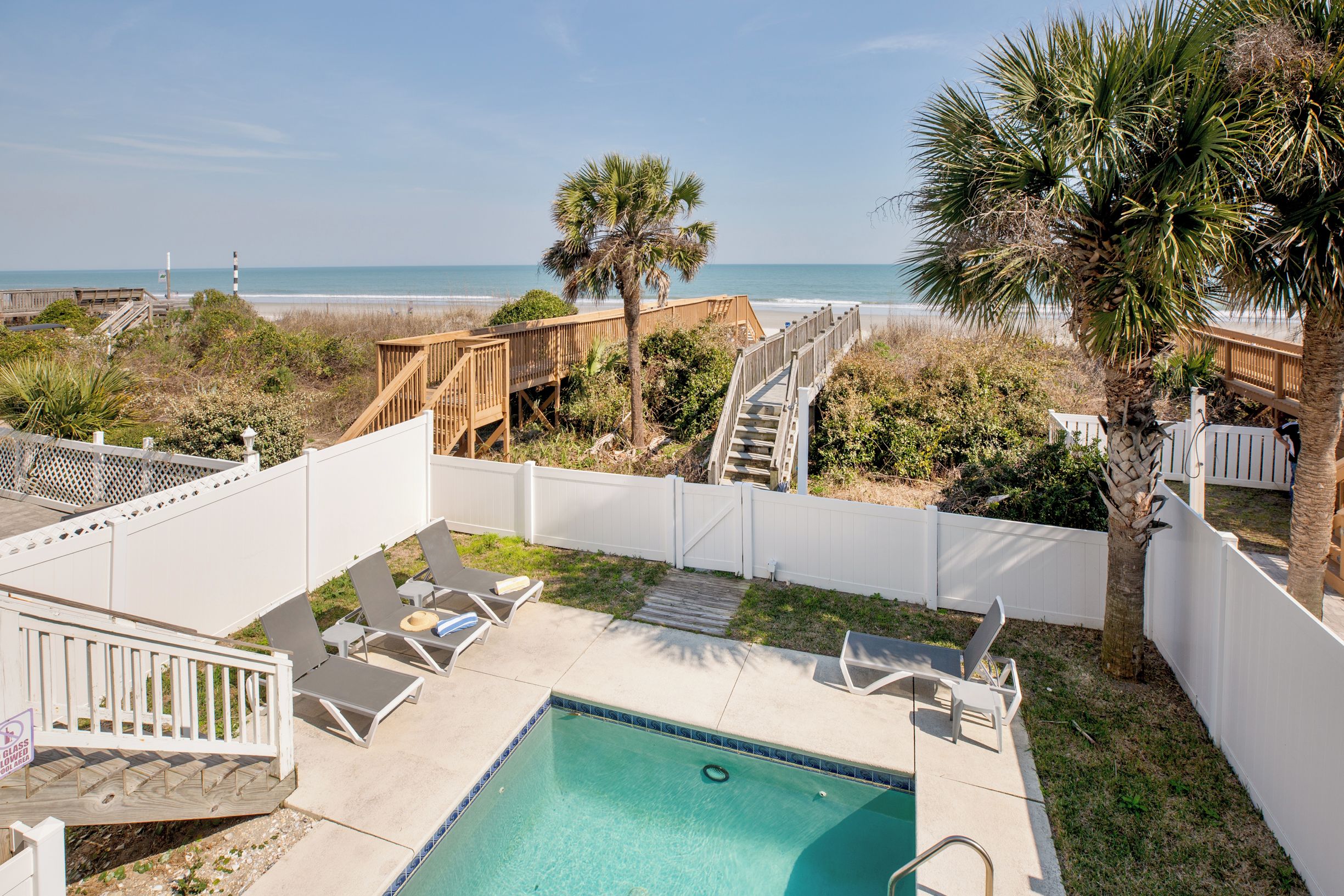 A backyard with a pool, lounge chairs, a wooden deck, and a view of the beach.