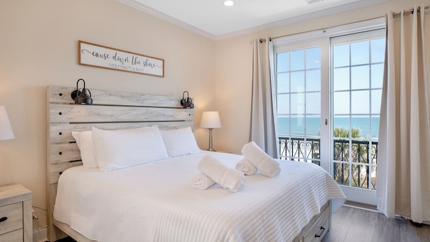 A hotel room with a bed, a nightstand, a lamp, a window with curtains, and a view of the sea. - A Wave From It All: Oceanfront & Pool (Surfside Beach)