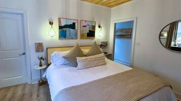 A bedroom with a wooden ceiling, a bed with white linens and patterned pillows, a small bedside table with a vase, and abstract wall art.