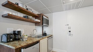 Una kitchenette compacta con microondas, tostadora, cafetera y un lavabo. Hay repisas de madera sobre la encimera con vajilla y vasos.