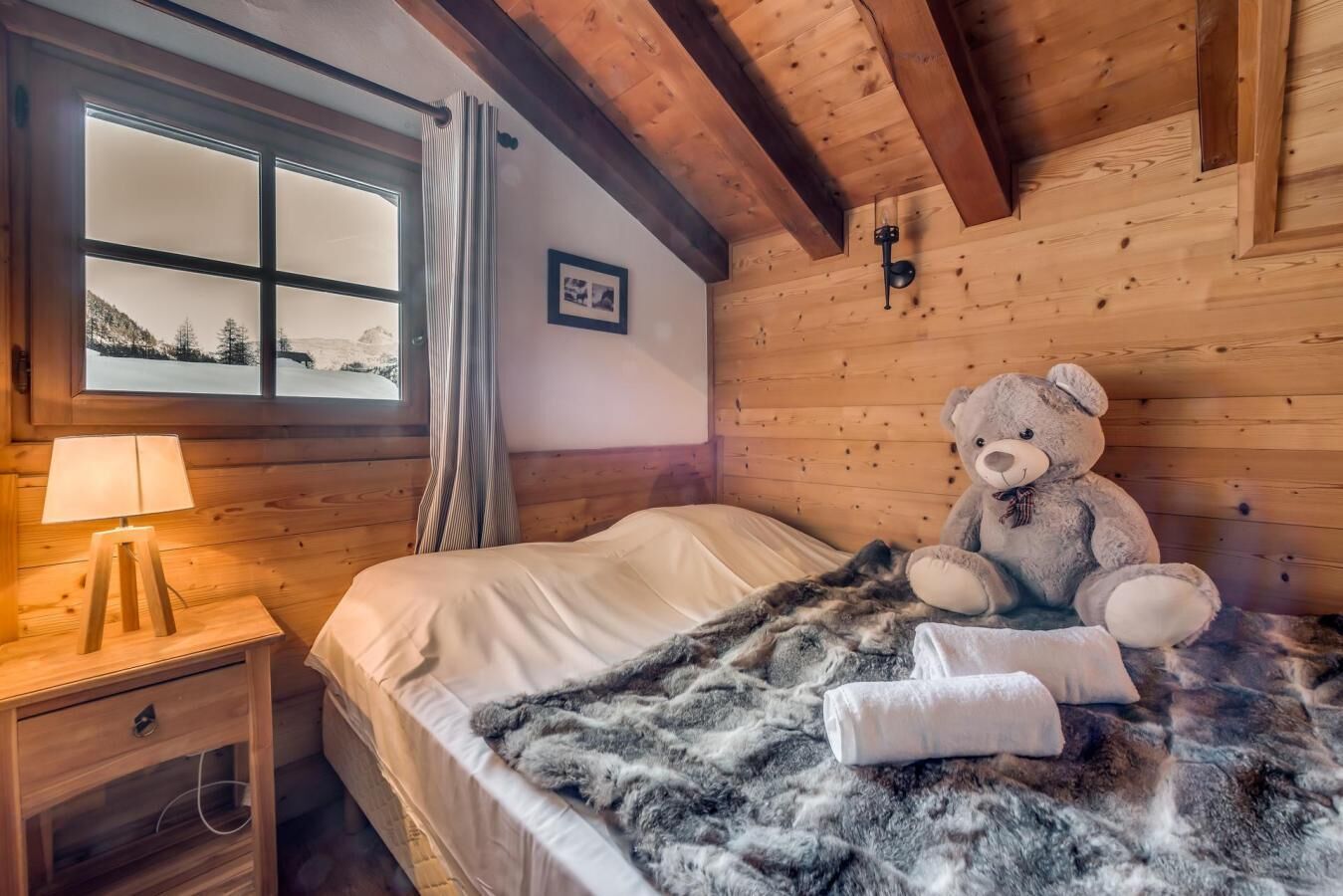 Una habitación acogedora con una cama, un osito de peluche, una lámpara y una ventana que da a un paisaje nevado.