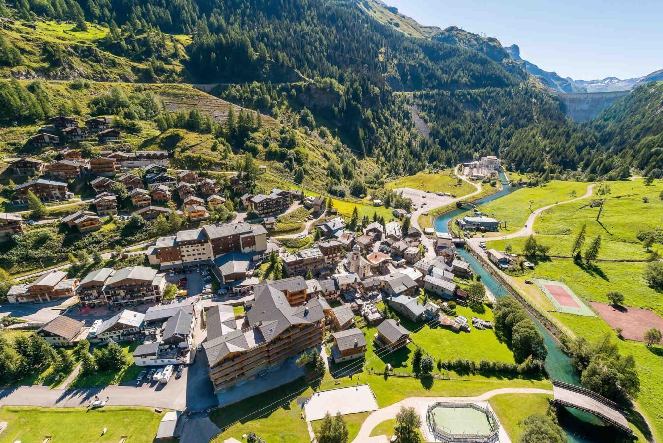 An aerial view of a mountain village with traditional chalet-style buildings, a river, and a bridge.