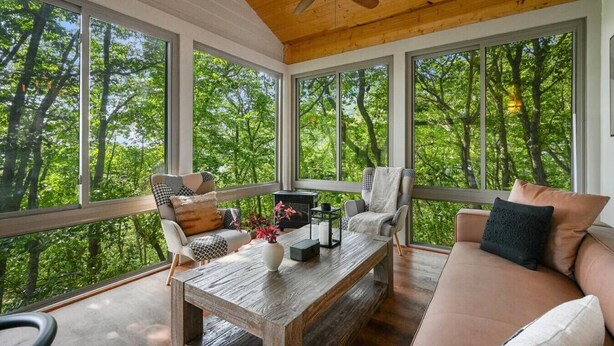 Amplia habitación con grandes ventanales que ofrecen vistas a un bosque. Cuenta con un ventilador de techo, una mesa de centro de madera y un sofá color beige.