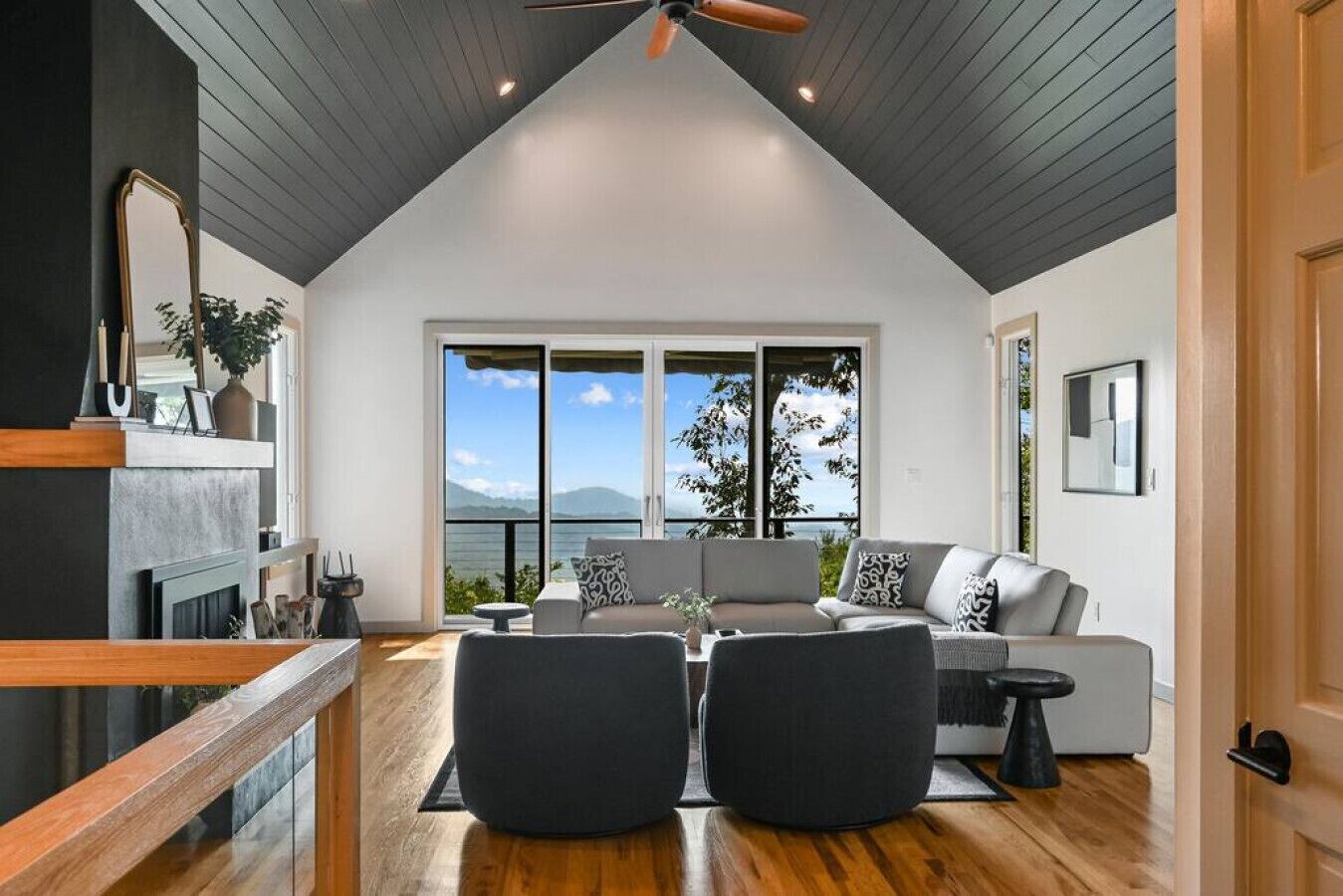 Un salón amplio con techo alto, un ventanal que ofrece vistas al exterior, un sofá gris y suelo de madera.