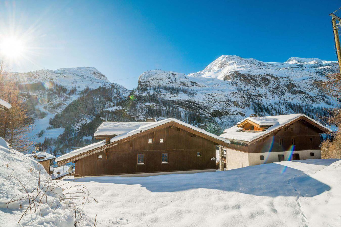 A wooden chalet with a sloped roof, surrounded by snow-covered mountains and clear skies.