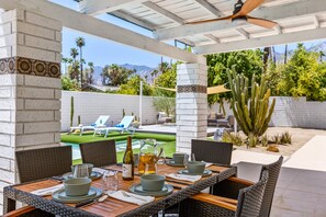 Une terrasse extérieure avec une table dressée pour le repas, entourée de chaises en osier, et offrant une vue sur une piscine et un espace verdoyant.