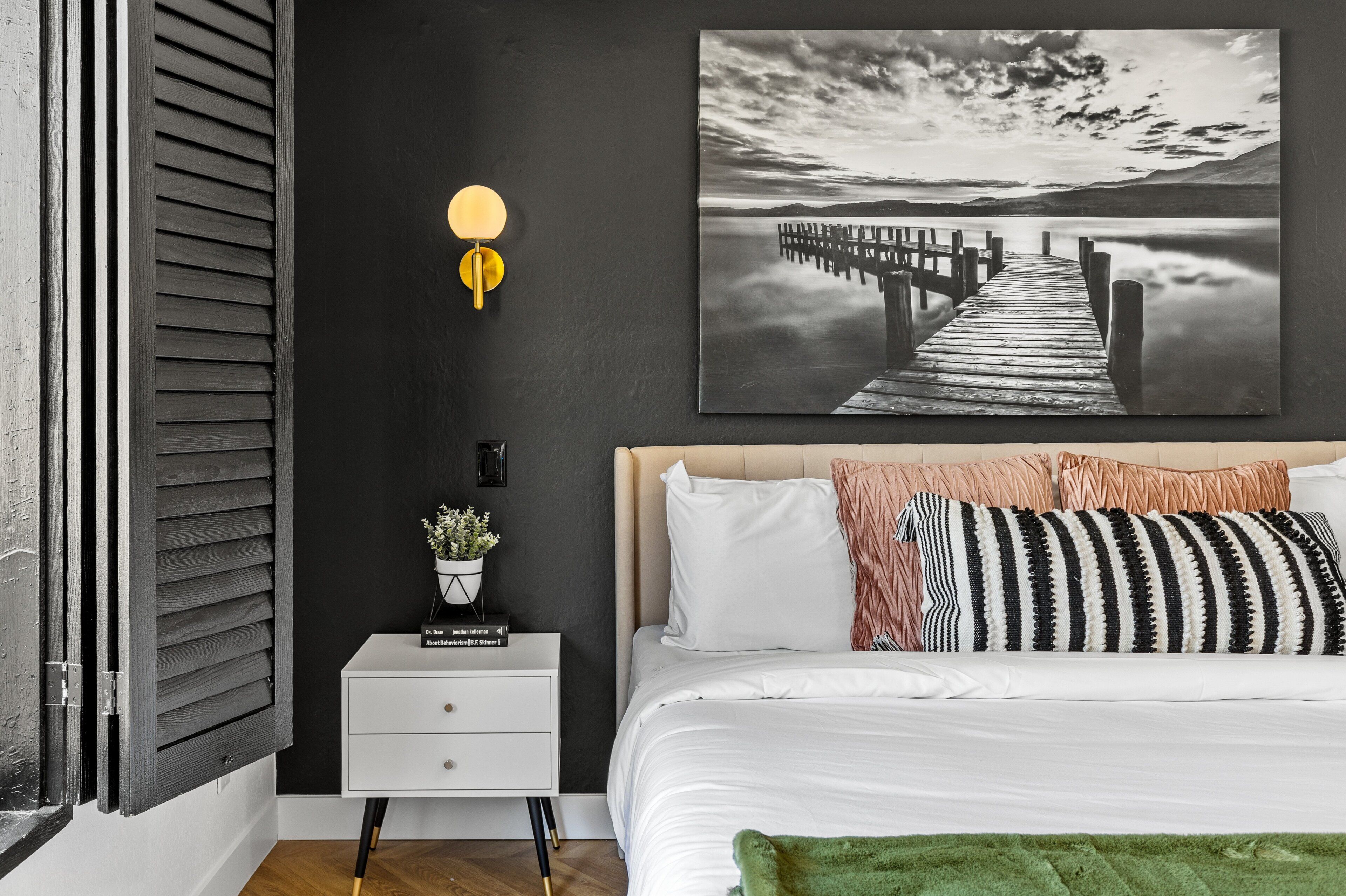 A bedroom with a bed, a nightstand, a wall-mounted lamp, a framed picture, and a window with blinds.