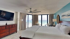Une chambre spacieuse avec un grand lit, un ventilateur de plafond, un téléviseur fixé au mur et une vue sur l’océan grâce à de grandes fenêtres.