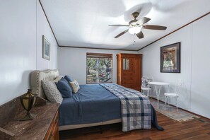 A bedroom with a bed, a ceiling fan, a wooden door, a small white table with chairs, and a window with a view of the outside.