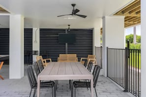 An outdoor patio with a wooden table and chairs, a ceiling fan, and a TV mounted on a black wall.