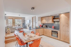 A modern kitchen with wooden cabinets, a dining area with orange chairs, and a living room with a sofa and a TV.