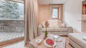 Ett mysigt vardagsrum med en stenmur, trägolv och ett stort fönster. Där finns en soffa med två nallebjörnar, ett matbord med en tallrik mat och glas, och en flaska vin.