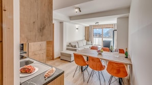Una cocina moderna con armarios de madera y encimera de mármol, un comedor con sillas color naranja y una mesa, y un espacio de sala acogedor con un sofá y vistas al exterior.