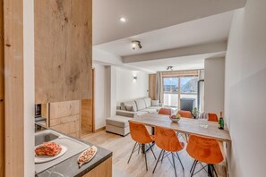 Una cocina moderna con armarios de madera y encimera de mármol, un comedor con sillas color naranja y una mesa, y un espacio de sala acogedor con un sofá y vistas al exterior.