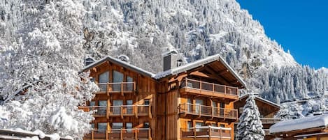 A wooden chalet with multiple balconies, surrounded by snow-covered trees and mountains.