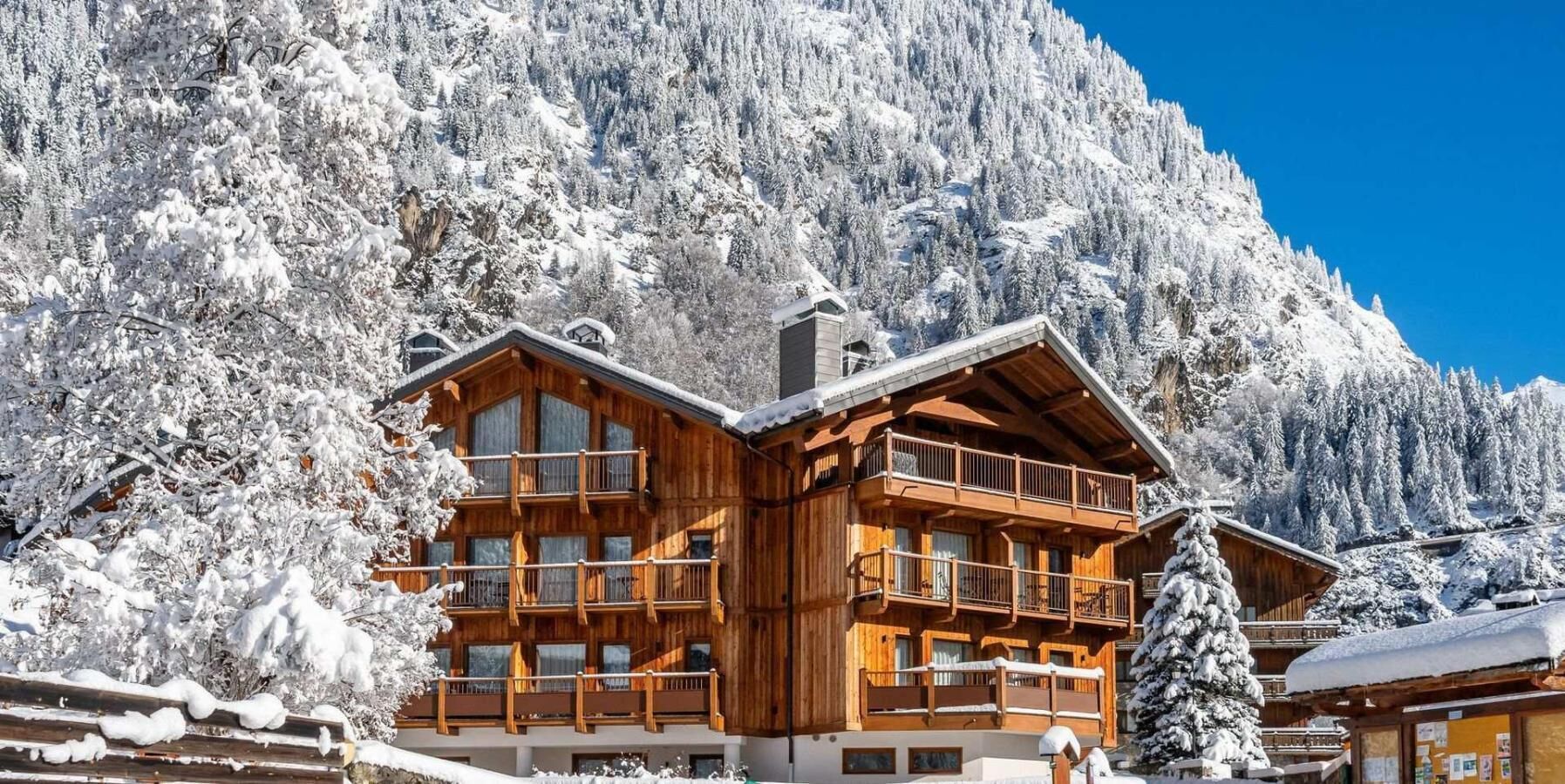 Una cabaña de madera con varios balcones, rodeada de árboles y montañas cubiertas de nieve.