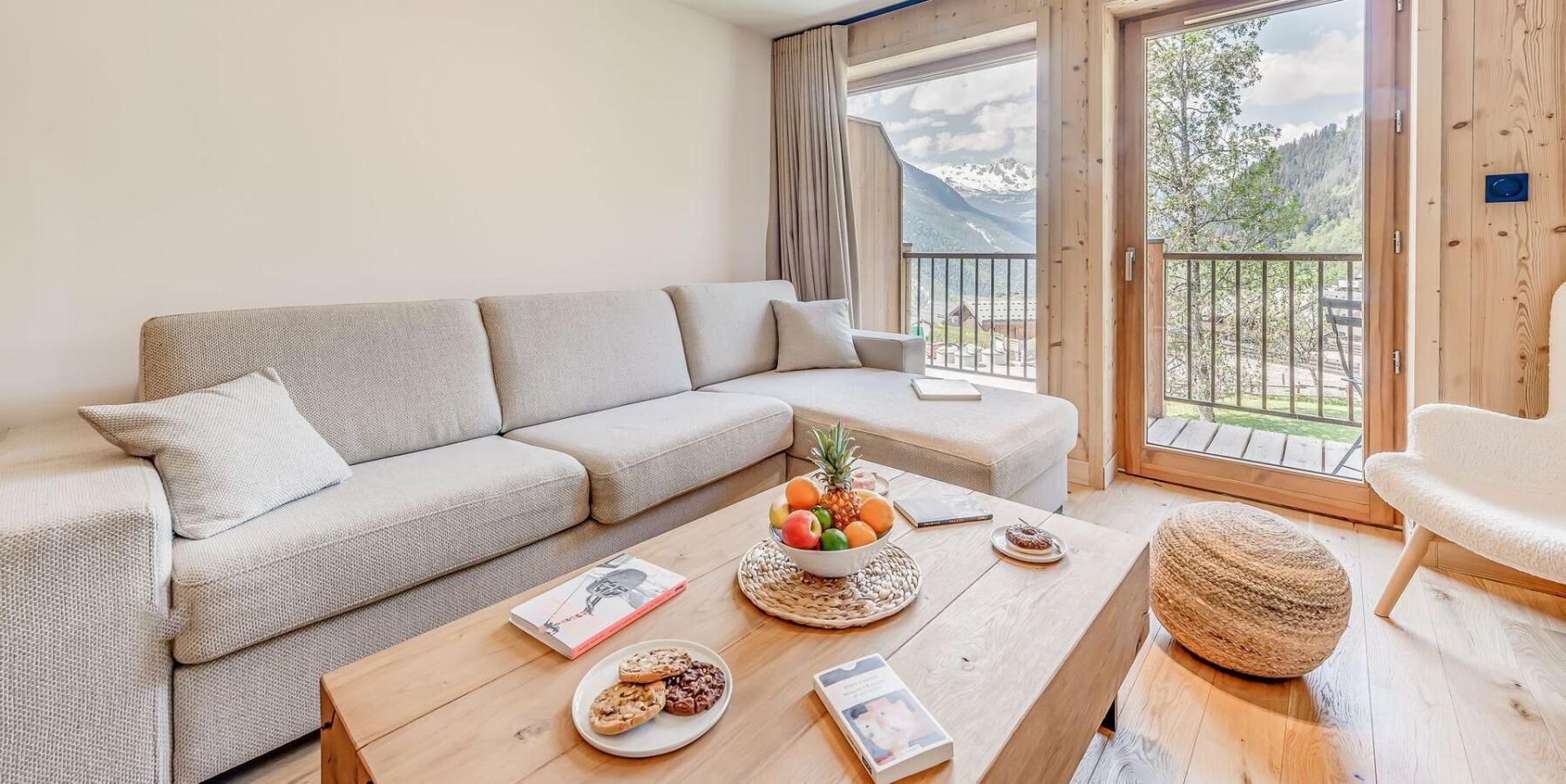 Ein gemütliches Wohnzimmer mit einer Couch, einem Holztisch mit Snacks und einem Buch sowie Blick auf Berge vom Balkon aus.