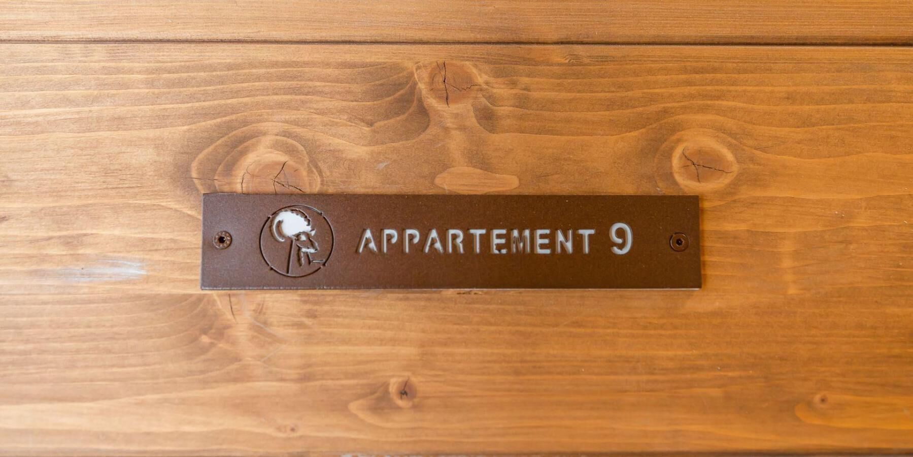 Een metalen plaquette met de inscriptie 'APPARTMENT 9', bevestigd aan een houten oppervlak.