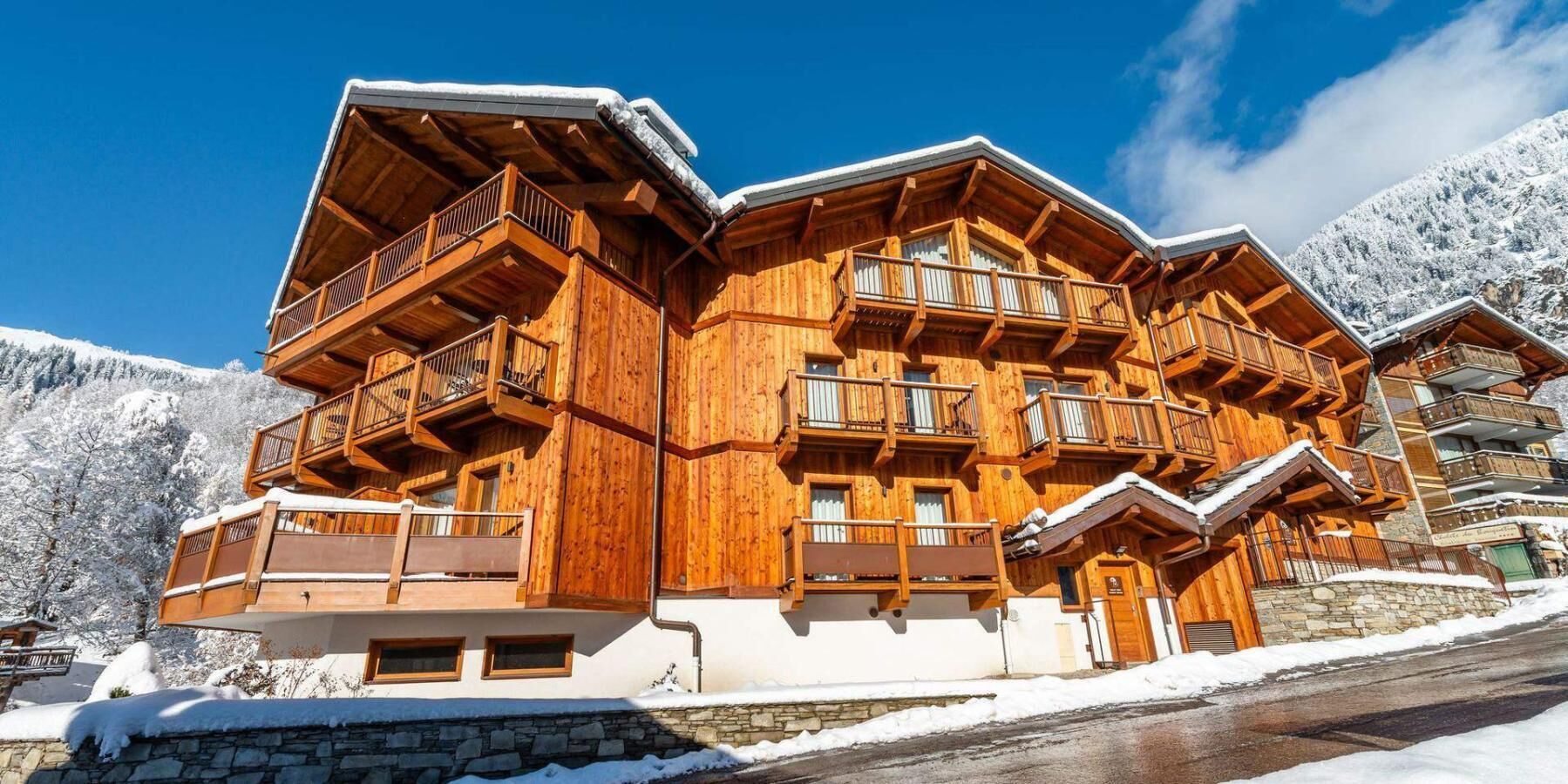 Un chalet en bois à plusieurs étages, doté de balcons, qui se dresse sur fond de montagnes enneigées.