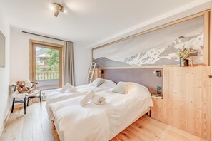Une chambre avec deux lits, une armoire en bois, un tableau représentant une montagne, un ours en peluche et une fenêtre donnant sur un paysage verdoyant.