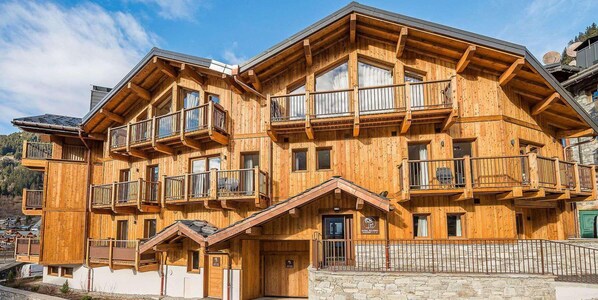 Un immeuble en bois à plusieurs étages, doté de balcons et d’une base en pierre, situé dans un cadre de station de ski.
