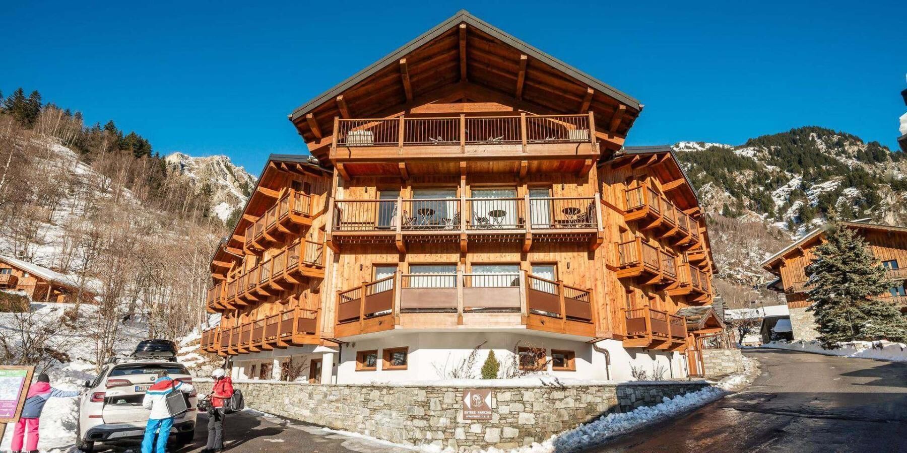 A multi-story wooden chalet with balconies, a stone foundation, and a ski resort setting.